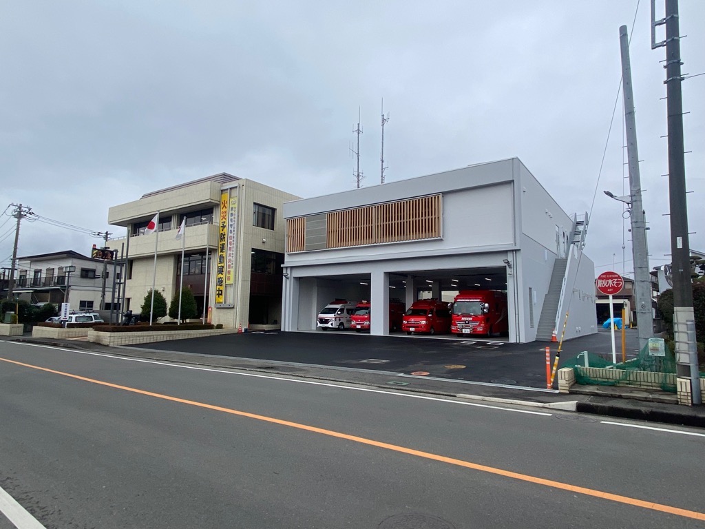 大通りに面して建つ、2階建ての古めの建物と、その右隣に真新しい白い建物があり、シャッターの開いた車庫には赤色の消防車が3台以上停車している消防本部の外観写真