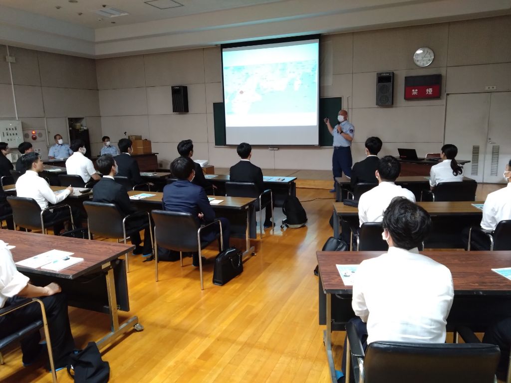 マスク姿の男性講師がスライドを投影しながら、多数の生徒または職員に向かって講義している様子を写した写真