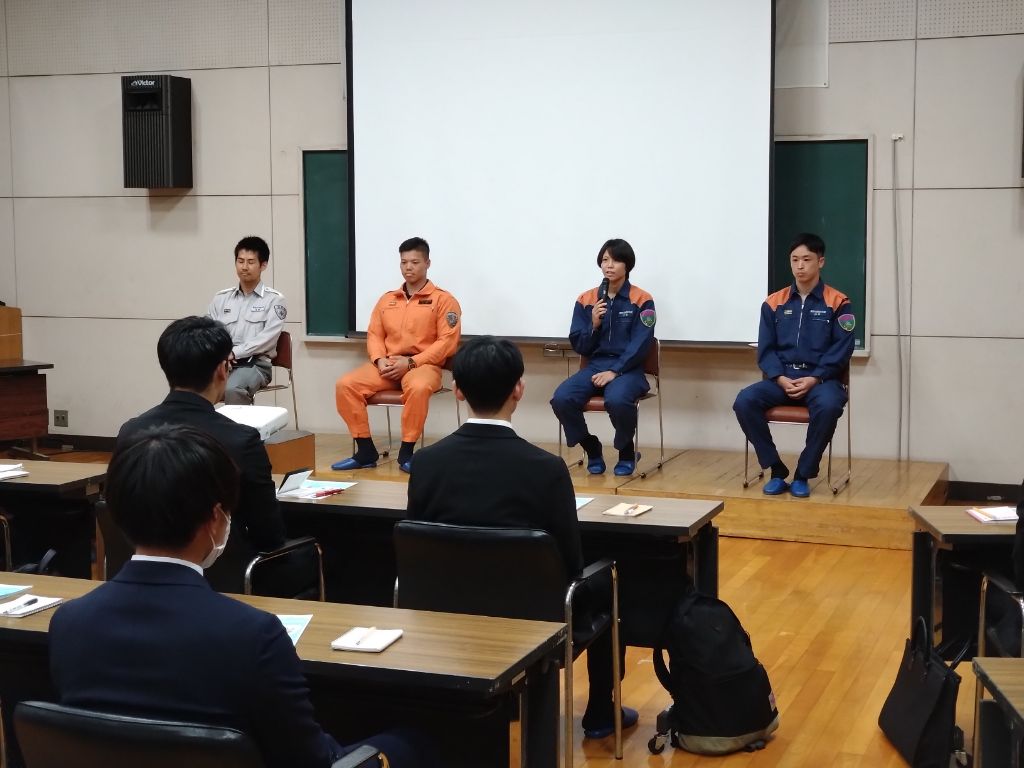 壇上に制服姿やオレンジ色のレスキュー服姿の男女4名が座り、座席に座っている数名の聴衆に向かって、中央の女性がマイクを持って話している様子の写真