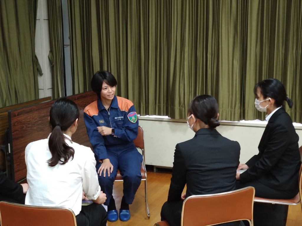 青い制服とオレンジ色のジャケットを着用した女性が、彼女を取り囲むように座っている3名のスーツ姿の女性と話している様子の写真