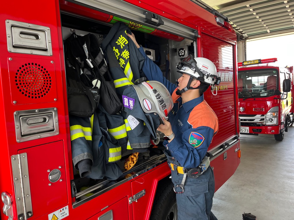 オレンジ色の防火服を着用した消防隊員が、赤い消防車の側面に開いた収納スペースから、ヘルメットを取り出し、吊り下げられている防火服を整頓している様子の写真