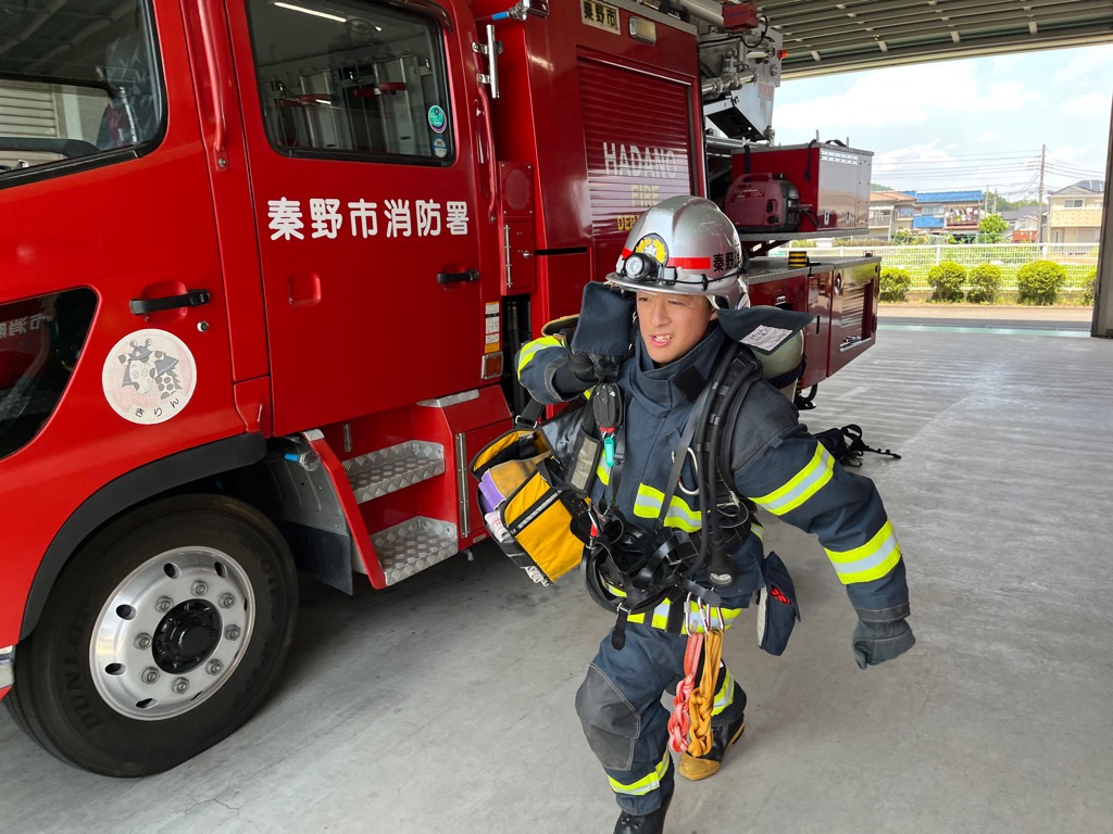 秦野市消防署と書かれた赤い消防車の横で、フル装備の防火服を着用し、ヘルメットをかぶり、背中にボンベやその他の装備を担いで前進している白石 純也さんの写真