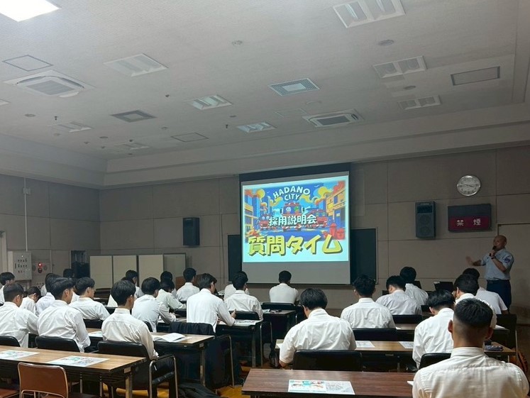 マスク姿の男性講師がスライドを投影しながら、多数の生徒または職員に向かって講義している様子を写した写真