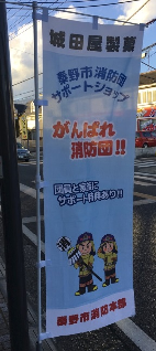 商店街の路上に、「がんばれ消防団!!」というメッセージとドット絵風のキャラクターが描かれた、秦野市消防団のサポートショップののぼり旗写真