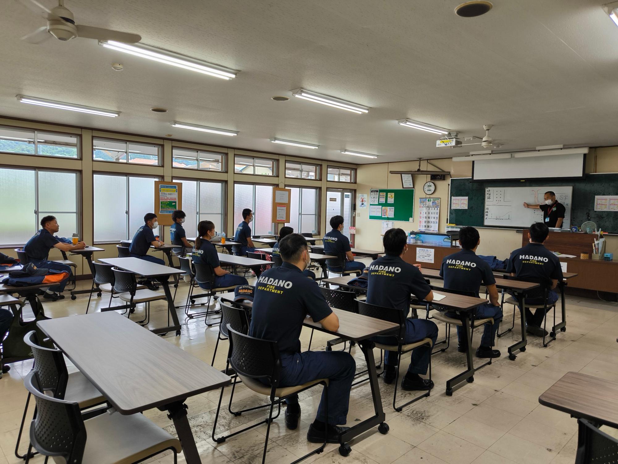 消防署員と思われる「HADANO DEPARTMENT」のロゴ入り制服を着た十数名が、教室でホワイトボードを使う講師の研修を真剣に受けている様子の写真