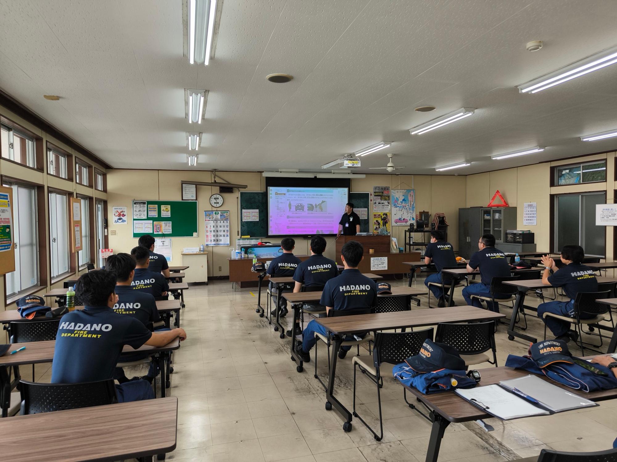 「HADANO FIRE DEPARTMENT」の制服を着た隊員たちが、教室でプロジェクターに映された画面を見ながら講師の研修を受けている風景の写真