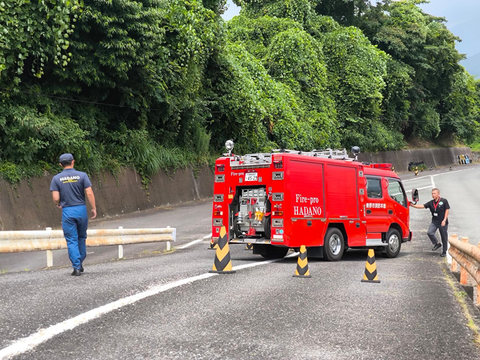 緑豊かな山道を背景に、秦野市消防本部の赤い消防車が停車し、隊員たちがコーンを置いて活動準備の指導を受けている様子の写真