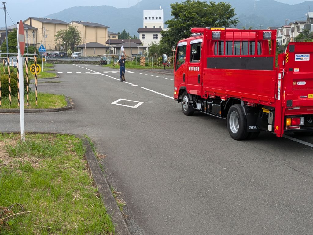 自動車教習所のようなコースで、秦野市消防本部の赤いトラックを、旗を持った隊員が後方に下がるよう誘導している訓練風景の写真