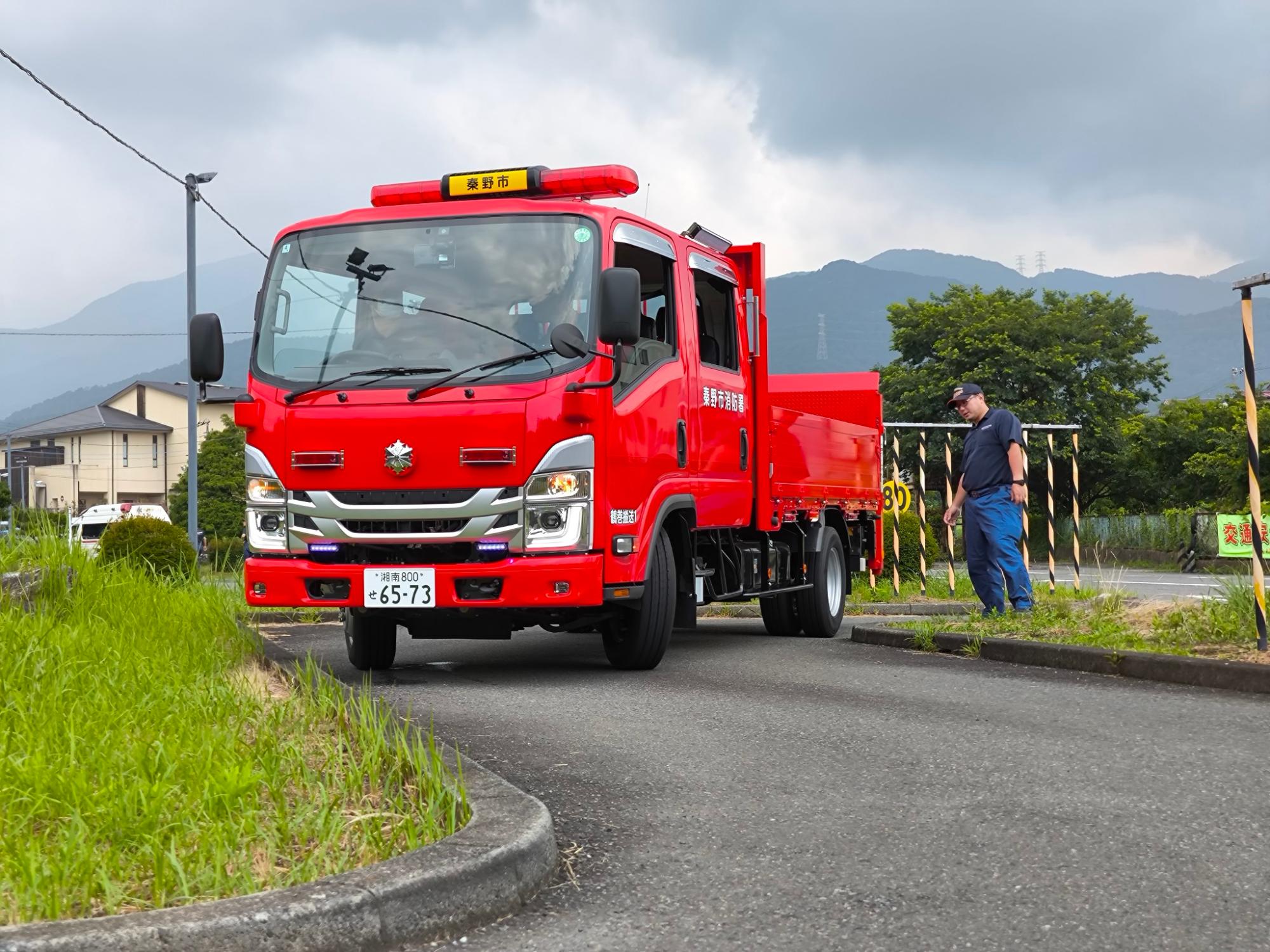 教習コースの狭いカーブを秦野市消防本部の赤いトラックがクランク走行し、その様子を隊員が見守っている写真