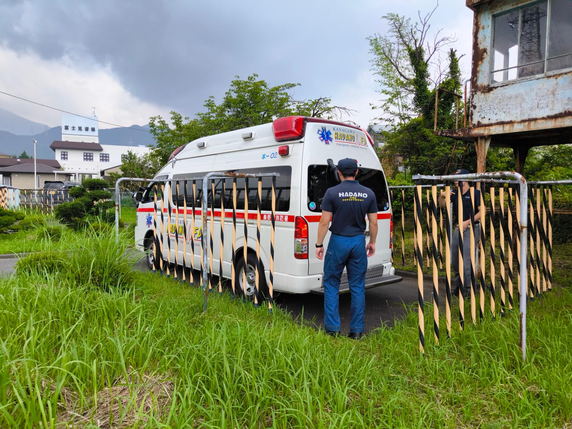 草が生い茂る空き地に停車した秦野市の救急車の後ろに、制服を着た隊員一人が立っている写真