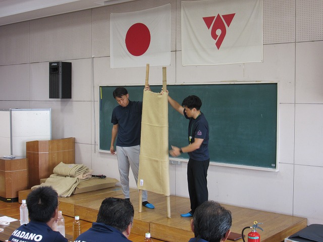 茶色の布製担架を2人の男性が床上のステージ上で実演しており、その様子を後部の聴衆が真剣な表情で見つめている訓練風景を捉えた写真