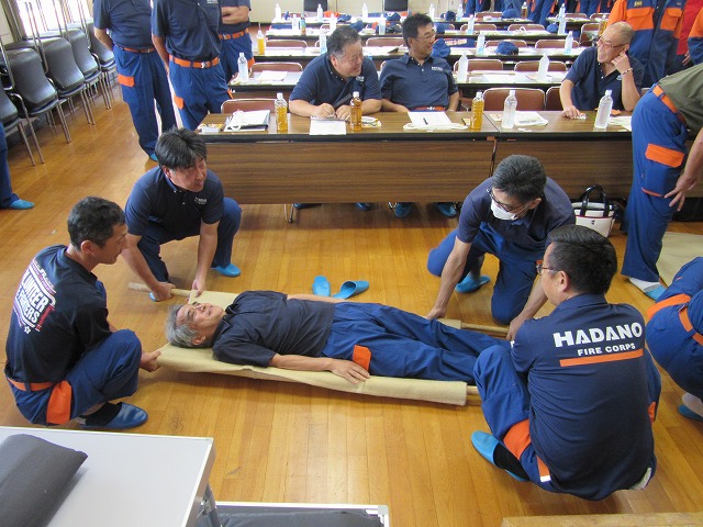 茶色の布製担架の上に横たわる男性を、4人の訓練参加者が棒を使って移動させる手順を訓練する様子の写真