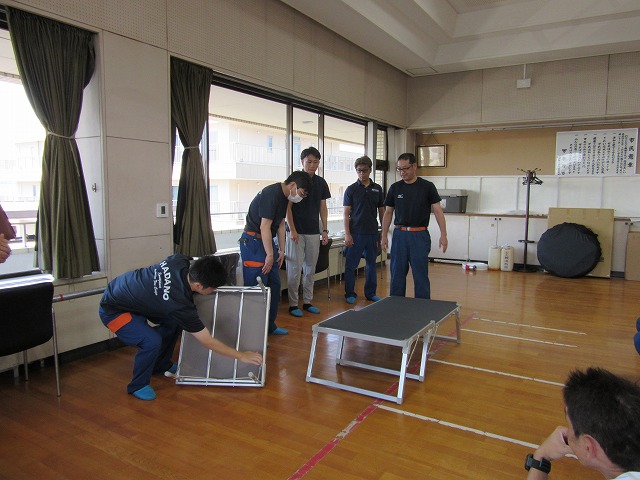 数人の男性が黒い布地の簡易的なベッドを組み立てており、1人がフレームの確認をしている訓練の様子を収めた写真