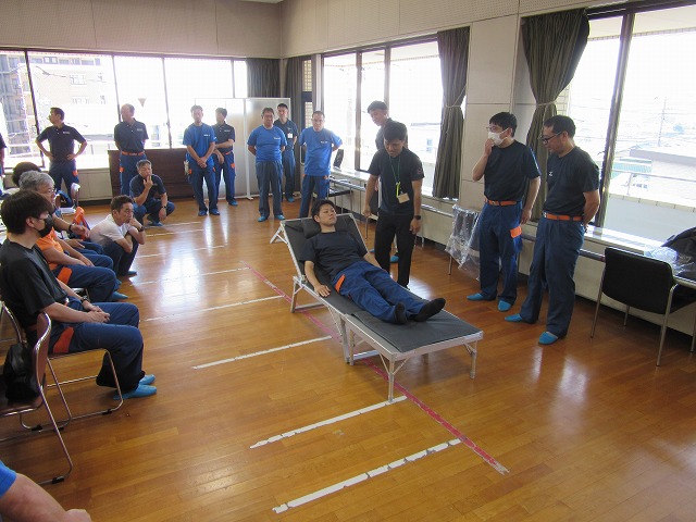 訓練の参加者たちが体育館のような広い室内に座ったり立ったりして見守る中、組み立てられた簡易ベッドに横たわる男性を指導者が確認しながら手順を説明している様子を捉えた写真