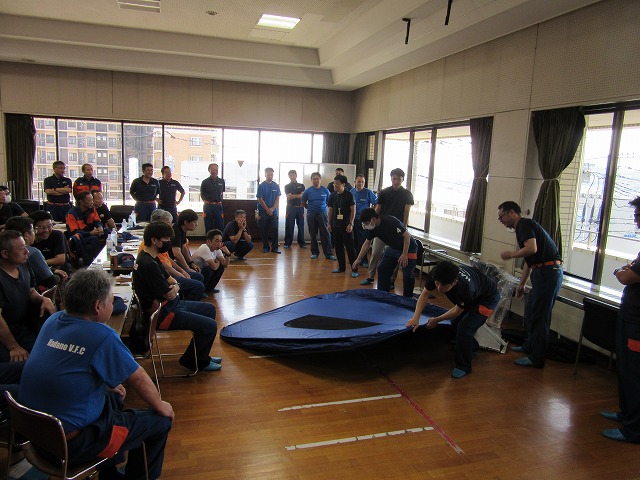 体育館のような広間の中で、多くの訓練参加者が見守る中、数人の男性が濃紺色の簡易テントを広げ、その使い方について実演している様子を捉えた写真