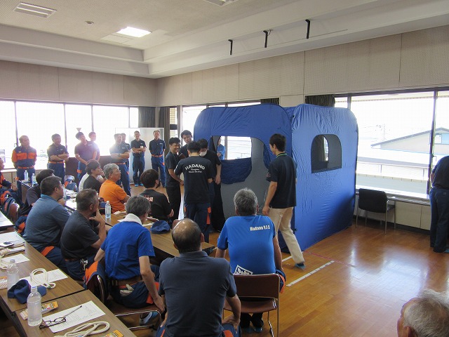 大勢の参加者が長机に座って見守る中、部屋の奥で数人の男性が濃紺の簡易テントのような救助用具を組み立てており、その様子を後方の関係者も注視している訓練風景を捉えた写真