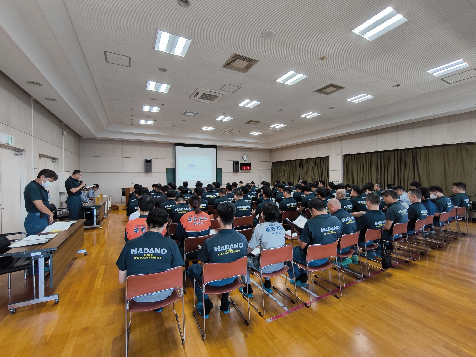 HADANO DEPARTMENTのロゴ入りTシャツを着用した男性らが、広い会場で演台の講師に背を向け、正面のスクリーンを見つめながら講義を聴いている講習会の写真