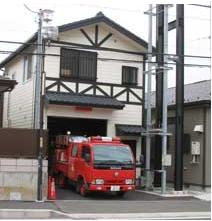 赤と白のツートンカラーの消防車が、出動に備えて1階のシャッターを開けた切妻屋根と白壁に黒の木組みが特徴的な消防団の車庫から前方に停車している外観写真