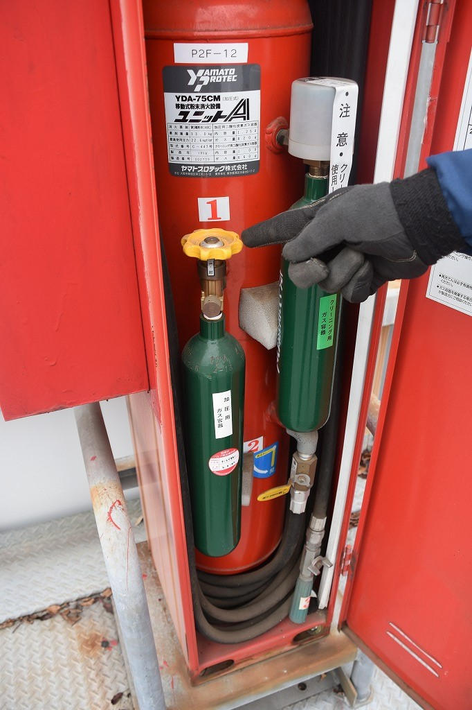 赤い移動式粉末消火設備の内部で、大きな赤いタンクの横にある緑色の加圧用ガスボンベの黄色いハンドルを指さしている写真