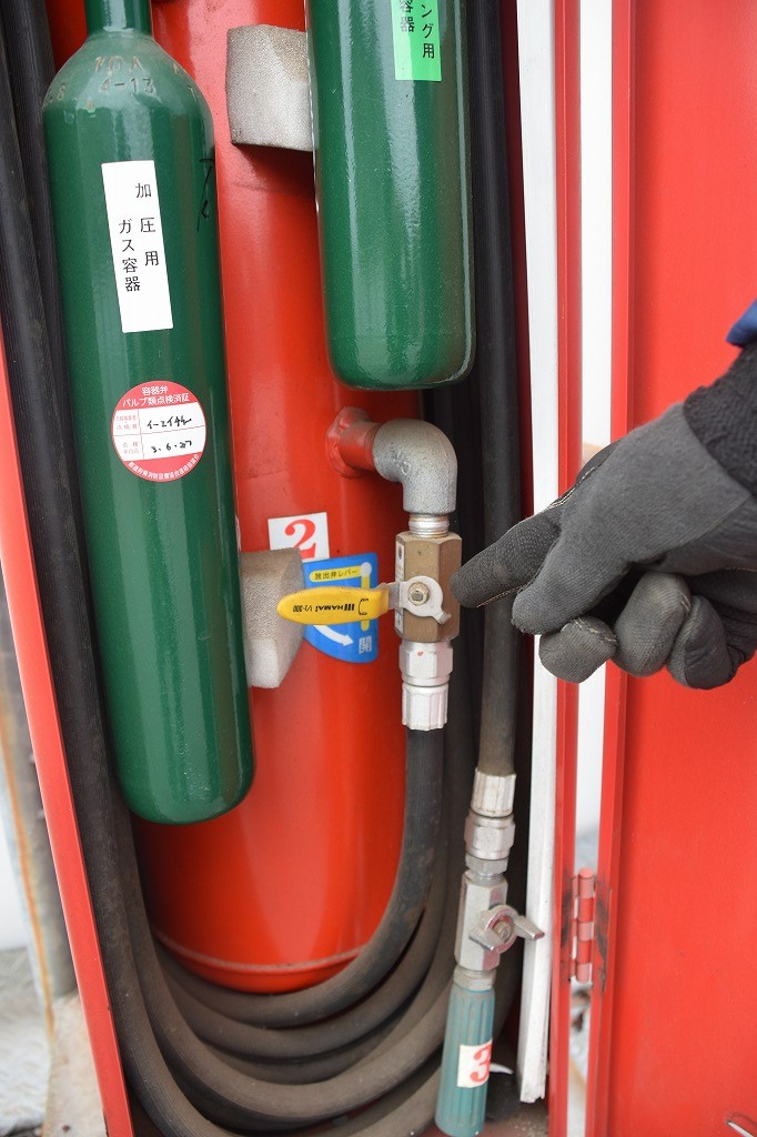 赤い移動式粉末消火設備の内部で、大きな赤いタンクと緑の加圧用ガス容器の間に設置された黄色いハンドルのバルブを、指さしている写真