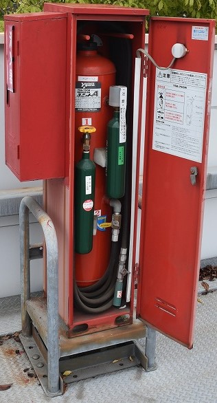 屋外に設置された「移動式粉末消火設備」の赤い格納箱の扉が開かれ、内部にある大きな赤い消火剤タンク、2本の緑色の加圧用ガスボンベ、黒いホースが見える写真