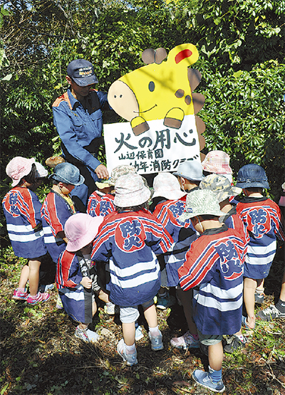 森の空き地で「火の用心」と書かれたキリンの看板を背景に、消防団員と思われる制服の男性が「放火」の文字が描かれた半纏を着た数名の園児たちに説明する様子の写真