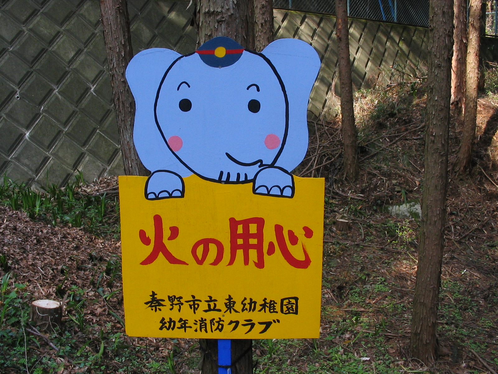 帽子をかぶった青い象のキャラクターが、黄色い看板に書かれた赤い太文字の「火の用心」と、「秦野市立東幼稚園 幼年消防クラブ」の文字を掲げている、山林内の注意喚起標識の写真