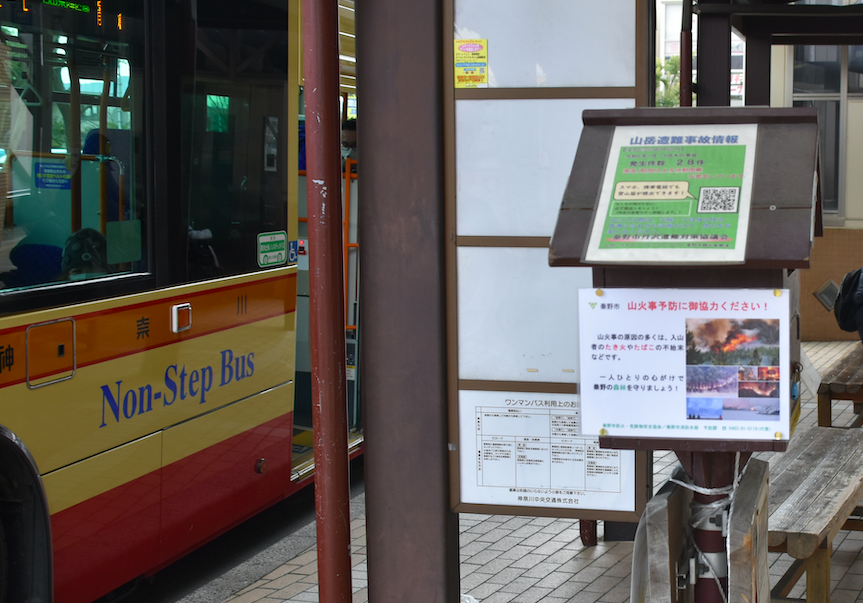 バス停のポールに掲示された「山火事予防に御協力ください！」という注意喚起のポスターの横に、黄土色と赤色の「Non-Step Bus」と書かれた路線バスが停車している光景の写真