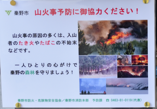「秦野市 山火事予防に御協力ください！」と書かれたポスターで、山火事の悲惨な様子の写真が複数コラージュされ、「山火事の原因の多くは、入山者のたき火やたばこの不始末などです」と注意を促している掲示物の写真