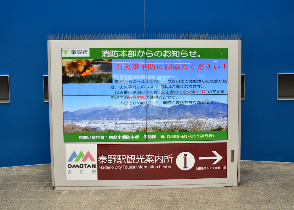青い壁の前に設置された大型デジタルサイネージに、「秦野市 消防本部からのお知らせ。山火事予防に御協力ください！」という警告文と、炎上する山火事、雪のない丹沢山地の遠景、そして秦野駅観光案内所の案内が表示されている掲示板の写真