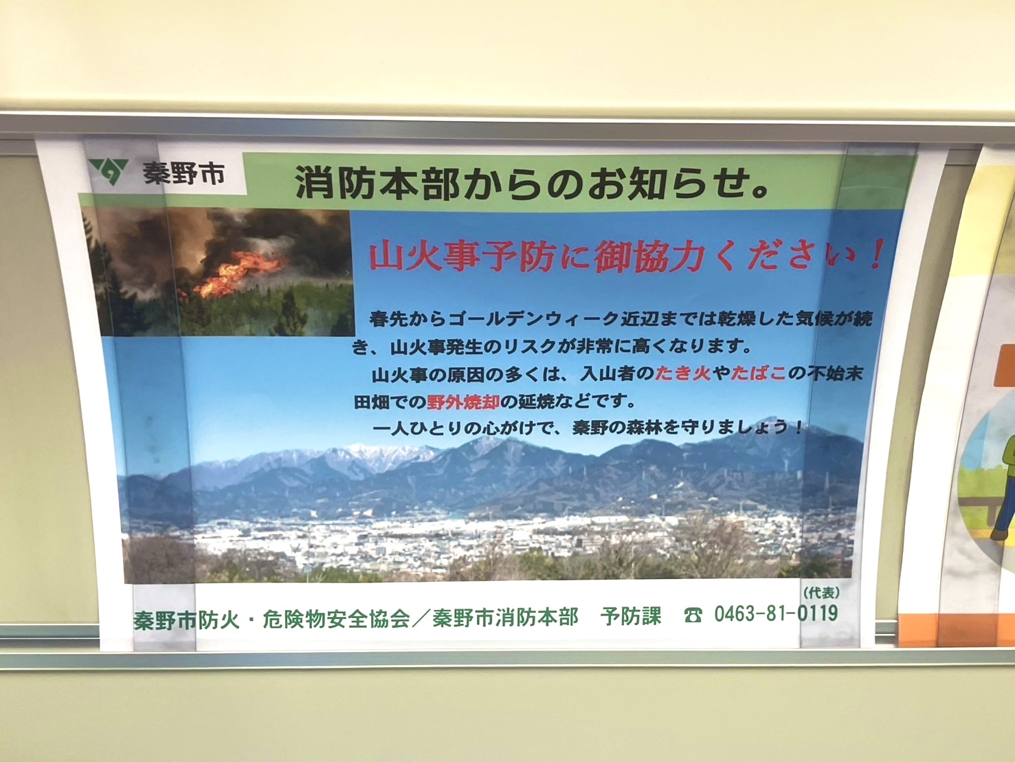 淡い黄色の壁に掲示されたポスターで、秦野市消防本部から「山火事予防に御協力ください！」と呼びかけがあり、山火事の写真と丹沢の山並みの写真が添えられている注意喚起の掲示物の写真
