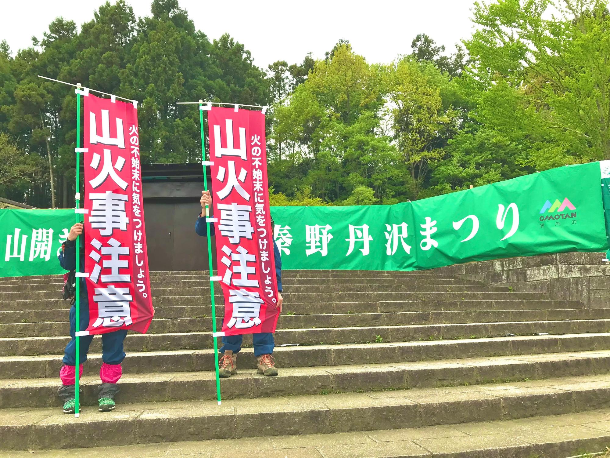 広場の石段前に立つ2名が、「山火事注意」と赤く大きく書かれた幟を掲げ、背景には緑色の「秦野丹沢まつり」と書かれた横断幕が見える啓発活動の様子の写真