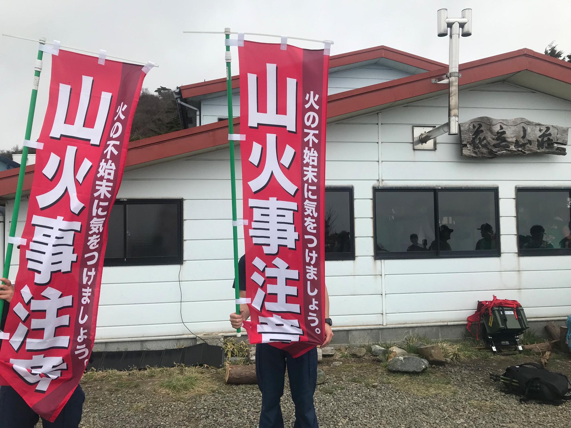 窓ガラスに人影が映る山小屋のような白い建物の前で、2名が「山火事注意 火の不始末に気をつけましょう。」と白抜き文字で書かれた赤い幟旗を掲げている啓発活動の様子の写真