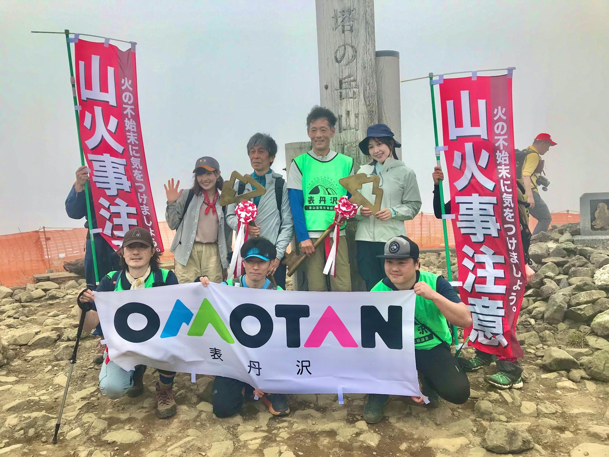 雲のかかった岩場の山頂で、登山者が「表丹沢」と書かれたロゴの横断幕を持ち、「山火事注意」の幟旗を両脇に立てて記念撮影をしている様子の写真