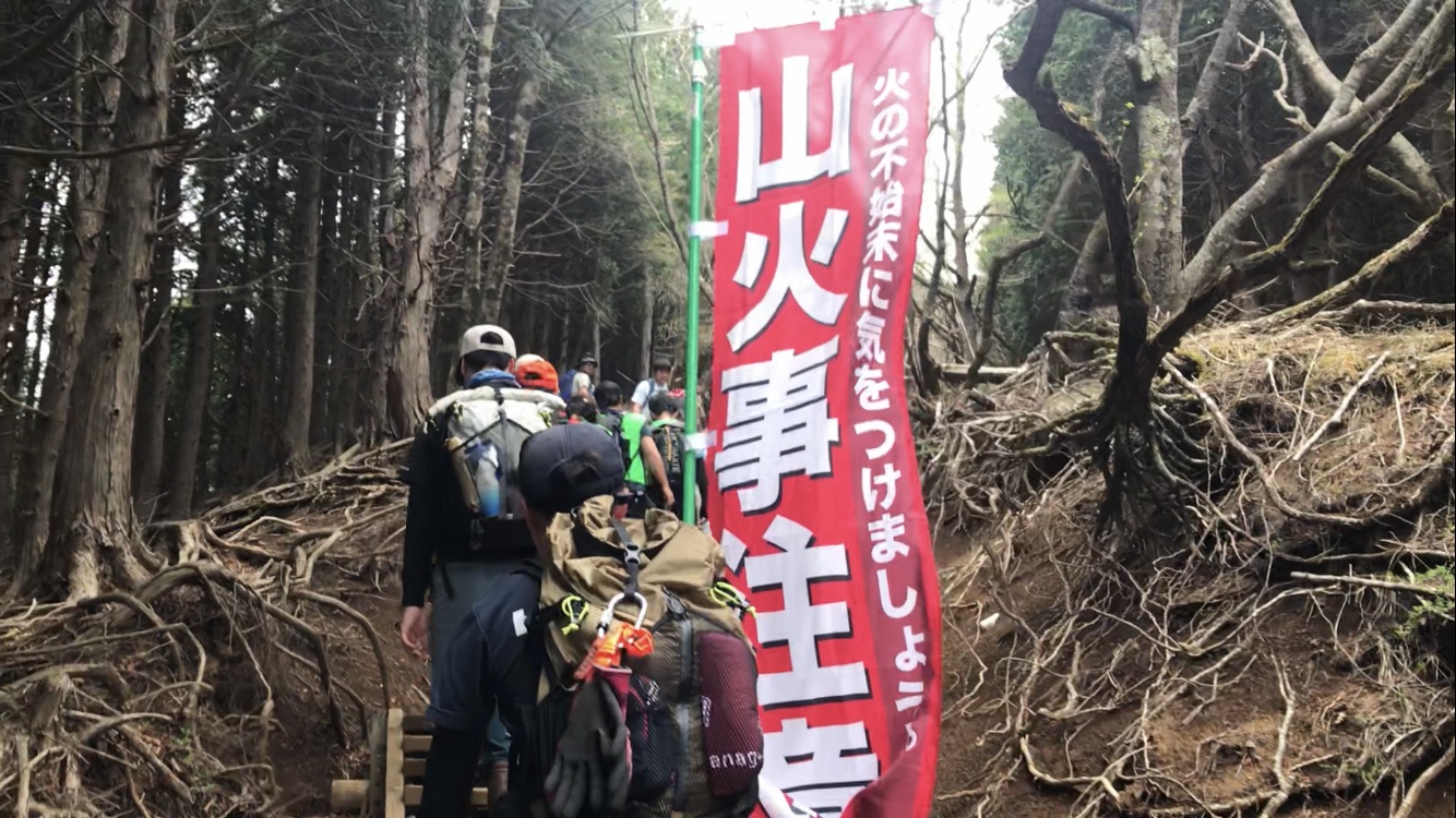 樹木の根がむき出しになった登山道で、赤い幟に書かれた「山火事注意」の文字の横を、リュックサックを背負った数名のハイカーが奥へと歩いていく、山登りの様子の写真