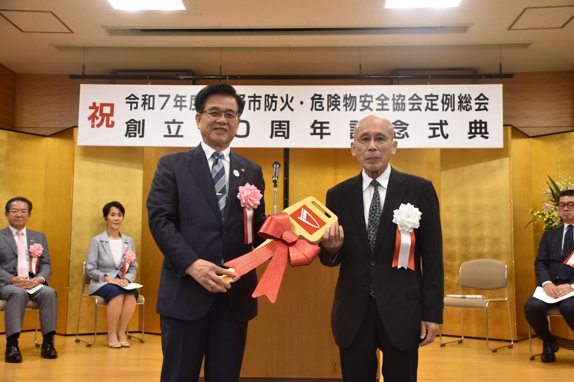 「祝 令和7年度 秦野市防火・危険物安全協会定例総会 創立50周年記念式典」と書かれた横断幕が掲げられている前でスーツ姿の市長がレプリカキーを、男性から両手で受け取っている記念写真