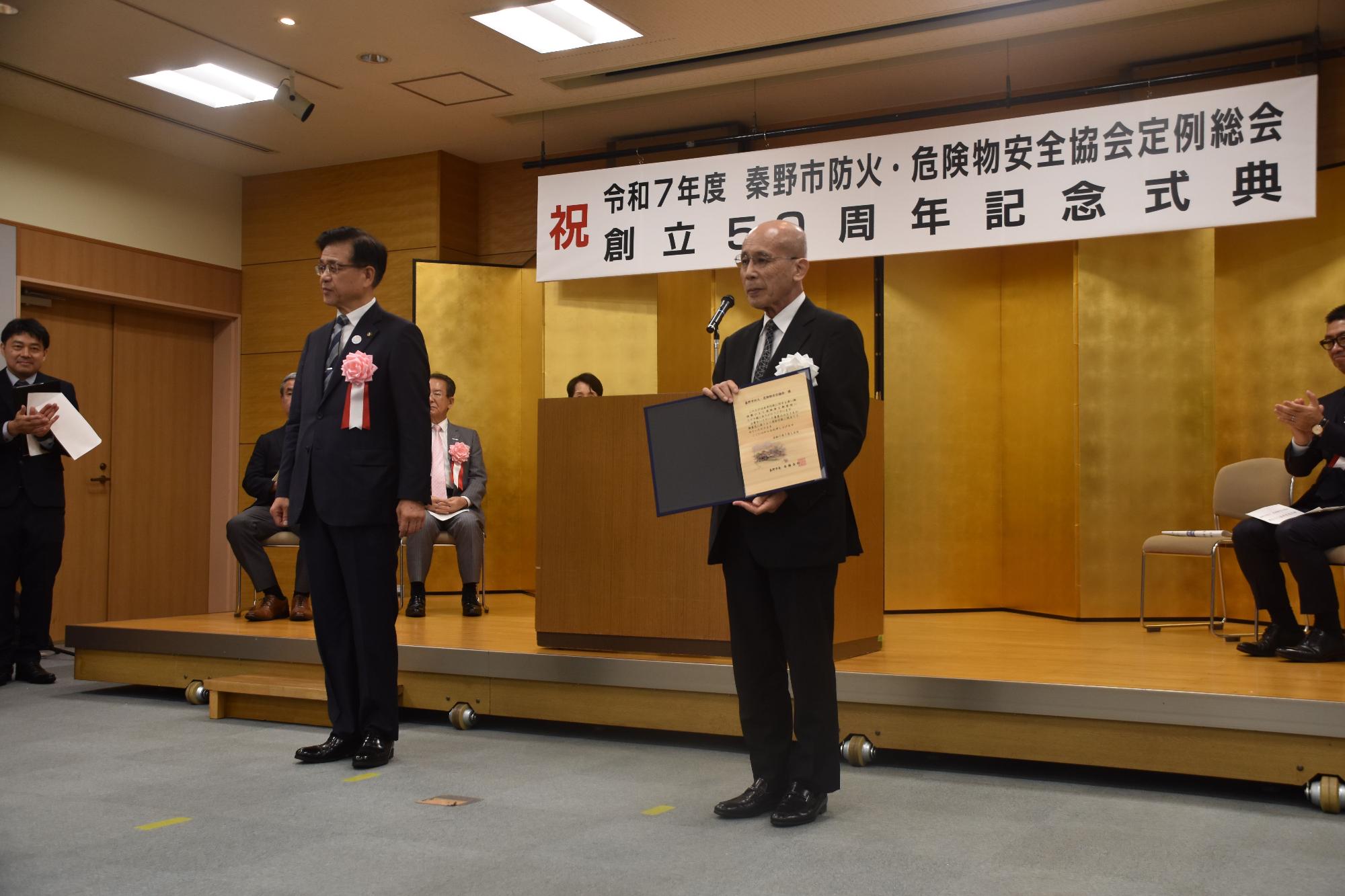 舞台上で行われている「令和7年度 秦野市防火・危険物安全協会定例総会 創立50周年記念式典」において、スーツ姿で立つ年配の男性が感謝状を両手に持って話をしている写真