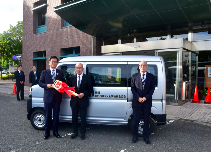 スーツ姿の市長と2人の男性が、車体に「寄贈 秦野市防火・危険物安全協会」と書かれてある銀色の軽自動車の前に立ち、市長と男性1人が大きな赤いリボンの付いたレプリカキーを持っている記念写真