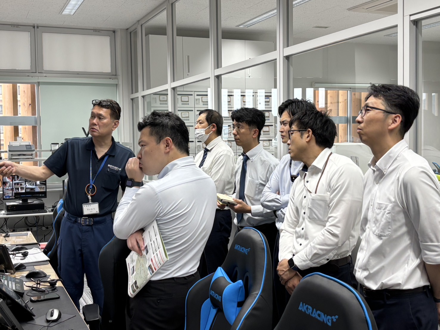 多くの男性職員が白いシャツやネクタイ姿で椅子の横に立ち、一人の濃紺のポロシャツ姿の男性がモニターを指差し、真剣な表情で指令室で説明を聞いている様子を捉えた写真