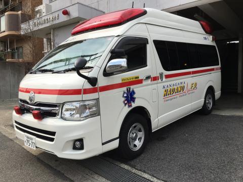 青い救急マークが車体に描かれ赤いラインが描かれた消防署本署の白い救急車が、建物の出入り口付近の舗装された場所に停まっている写真