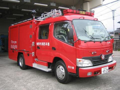 「消防署 HADANO」と英語と日本語で書かれた真っ赤な中型消防車で、車体側面には「秦野市消防本部」と書かれており、赤色灯と銀のハシゴが搭載された消防ポンプ自動車の写真