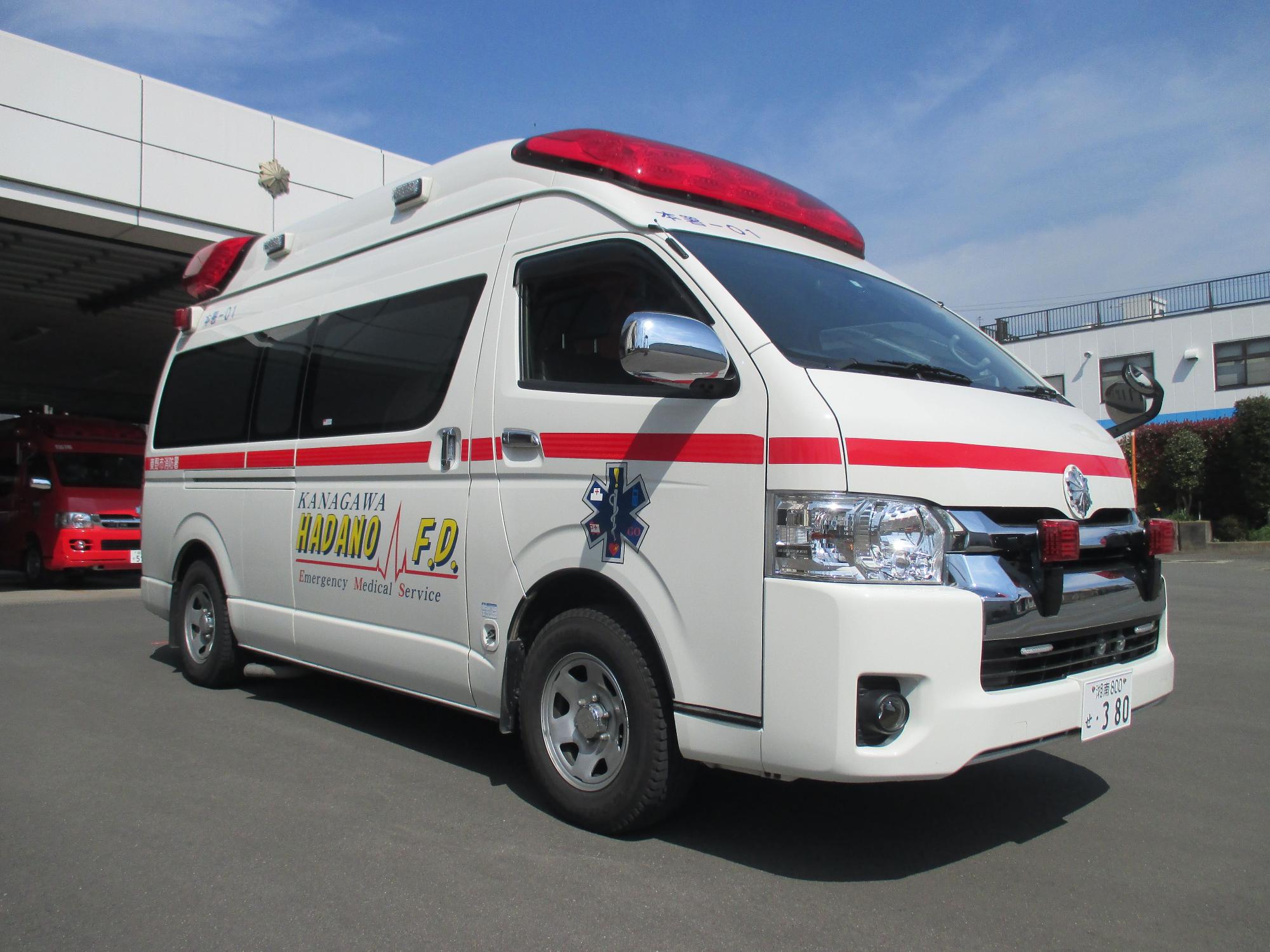 上部に赤色灯を設置した白いバンタイプの消防署本署の救急車で、車体には「KANAGAWA HADANO F.D. Emergency Medical Service」と書かれ、建物の前に停まっている写真