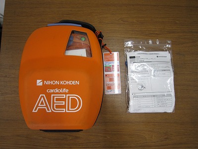 オレンジ色のケースに入った電気ショック用AEDと説明書らしき紙が並べて置かれている写真