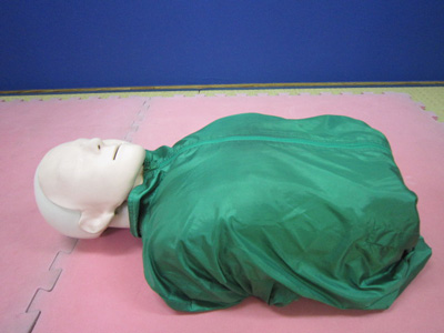 ピンク色のジョイントマットの上に、緑色のトレーニングウェアを着た救命講習用の白い顔をしたマネキンの上半身が、仰向けに置かれている心肺蘇生訓練人形（成人）の写真