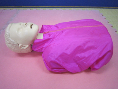 ピンク色のジョイントマットの上に、ピンク色のトレーニングウェアを着た救命講習用の白い顔をしたマネキンの上半身が、仰向けに置かれている心肺蘇生訓練人形（小児）の写真