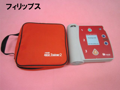 ピンクの背景の左上にフィリップスの文字があり、赤い収納ケースと、液晶画面や操作ボタンがついたAEDトレーナー2の本体が並べて置かれている訓練用AEDの写真