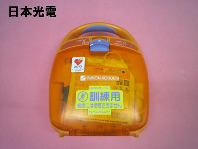 ピンクの背景の左上に日本光電の文字があり、オレンジ色のAEDトレーニングユニット本体には「訓練用」「救命には使用できません」と書かれた黄色いステッカーが貼られている訓練用AEDの写真