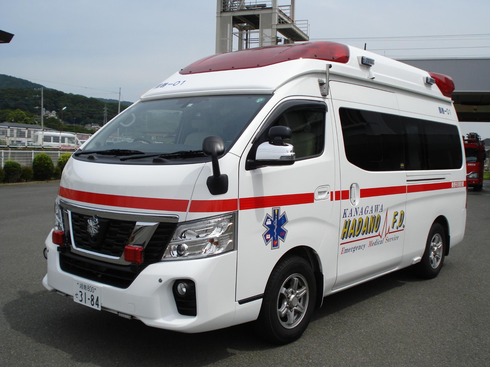 屋外の舗装された場所に停まっている、白い車体に赤いラインが入り青い六角形のマークが描かれ、上部に赤色灯を装備した消防署鶴巻分署の救急車の写真