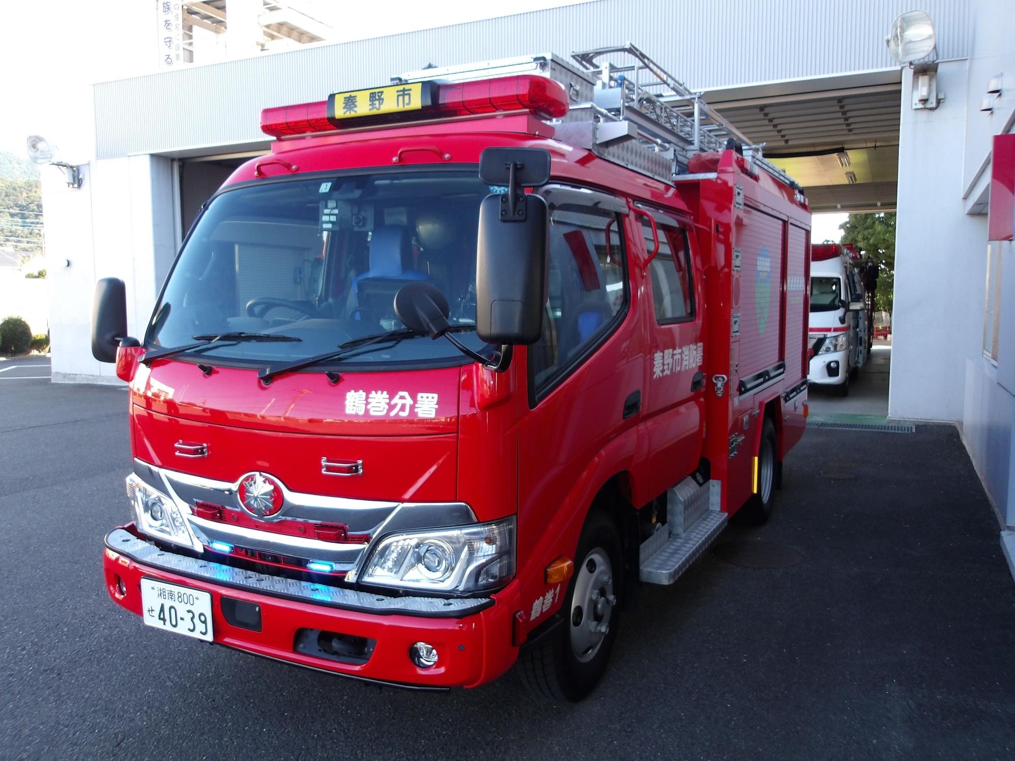 消防署の前に停車している真っ赤な中型消防車で、運転席の上に「秦野市」と表示され、車体側面に「鶴巻分署」「秦野市消防署」と記載されている消防ポンプ自動車の写真