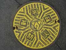 黒い円形のマンホール蓋で、蓋全体が鮮やかな黄色に塗られ、中央の消防章とそれを囲む放射状の線や、周囲に配置された「防」「火」「貯」「水」「槽」の漢字一文字が四角い枠に収まっているデザインの防火水槽の写真